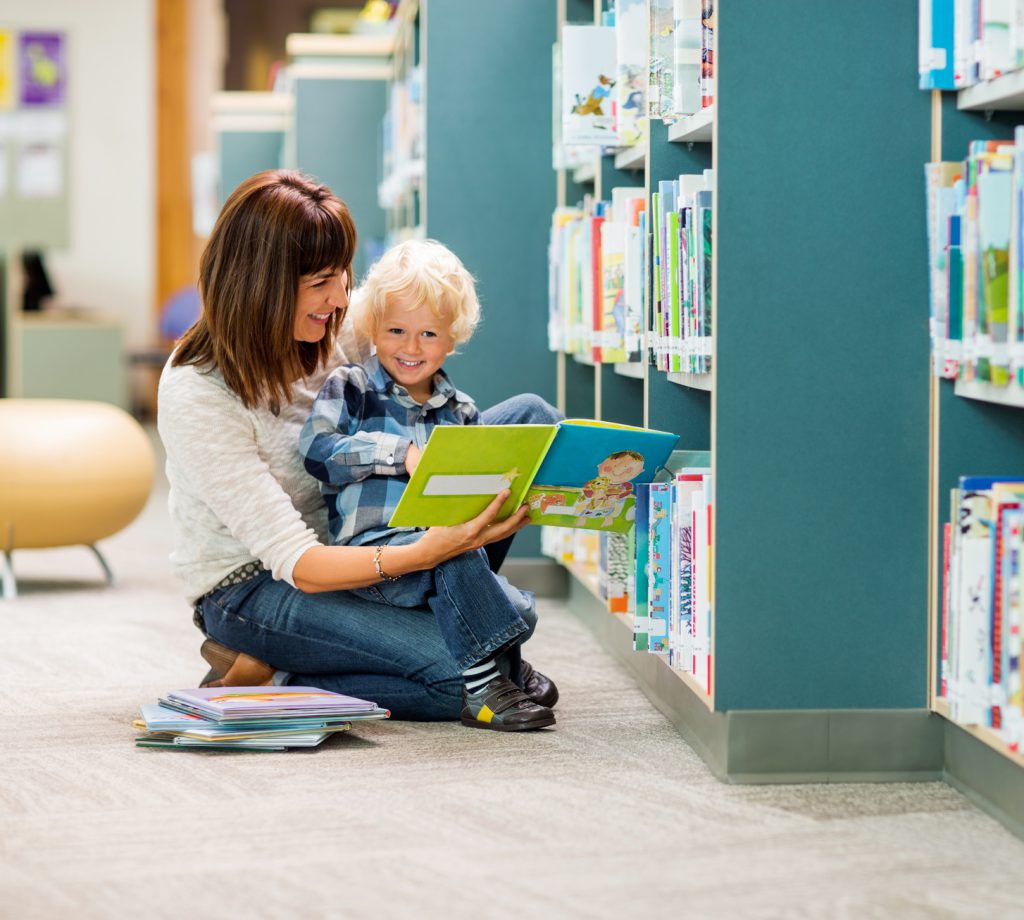 Full length of smiling boy and teacher reading book by bookshelf in library
